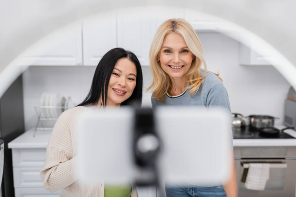 Blogueurs interraciaux joyeux regardant smartphone flou dans la lampe annulaire à la maison  - Photo, image