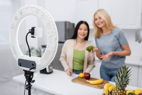 Cellulaire et lampe anneau près des amis interracial flous avec des fruits dans la cuisine  - Photo, image
