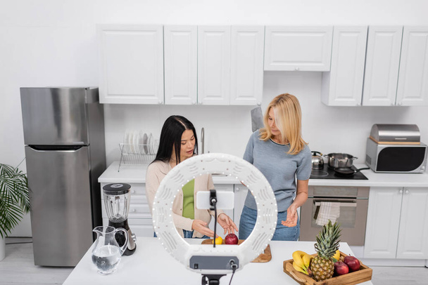 Amici multietnici che guardano frutta vicino smartphone e lampada ad anello in cucina  - Foto, immagini
