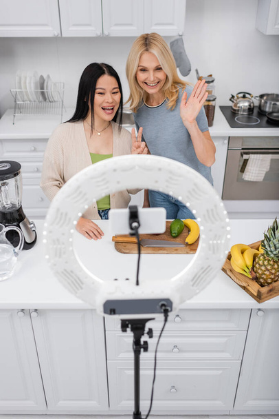 Bloggers interraciales positivos haciendo gestos en el smartphone en la lámpara de anillo cerca de frutas frescas en la cocina  - Foto, imagen