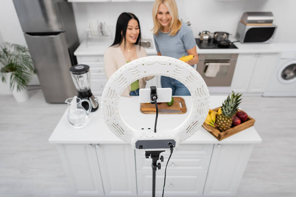 Мобильный телефон в лампе возле размытых многонациональных друзей с фруктами на кухне  - Фото, изображение
