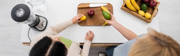 Vue aérienne des femmes tenant des fruits mûrs près de la planche à découper et du mélangeur dans la cuisine, bannière  - Photo, image