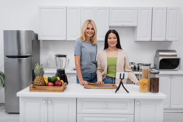 Cheerful interracial bloggers looking at camera near fruits and cellphone on tripod at home  - Photo, Image