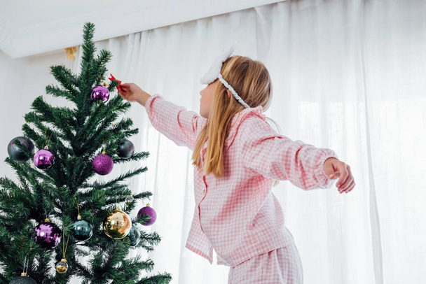 Petite fille en pyjama confortable décorant l'arbre de Noël. . Photo de haute qualité - Photo, image
