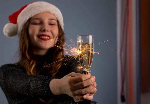 Atractiva chica alegre con el pelo rizado en el sombrero de Navidad sonriendo, la celebración de espumoso y beber champán, aislado sobre fondo gris
.. - Foto, Imagen