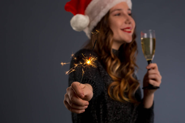 Привлекательная веселая девушка с вьющимися волосами в рождественской шляпе, улыбающаяся, держащая искру и пьющая шампанское, изолированная на сером фоне
.. - Фото, изображение