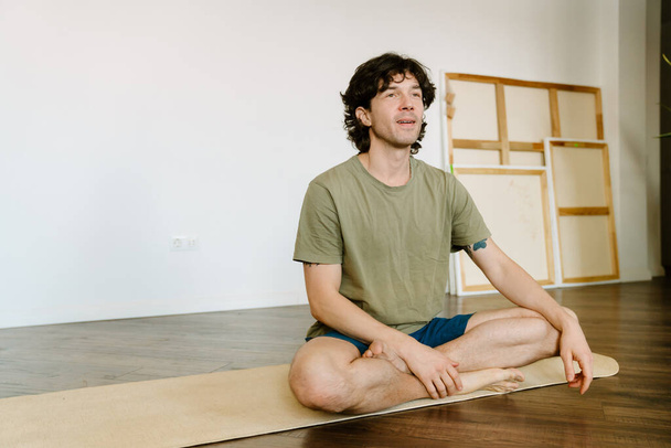 White man wearing shorts sitting on mat during yoga practice at home - Foto, Bild