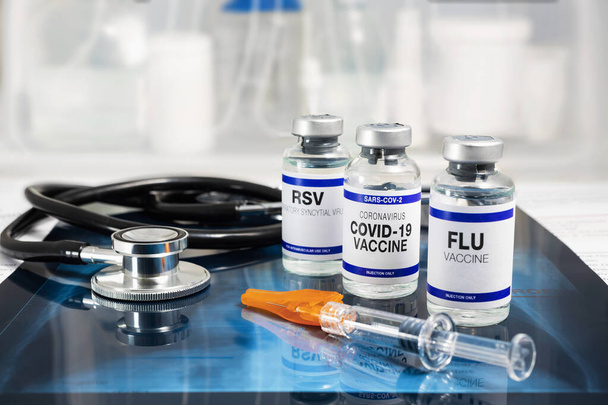 Flessen vaccin tegen influenza virus, respiratoir syncytieel virus en Covid-19 voor vaccinatie. Flu, RSV en Sars-cov-2 injectieflacons met Coronavirus vaccin boven Radiografie pulmonaal met stethoscoop - Foto, afbeelding