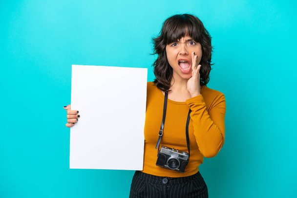 Молодая фотограф латинская женщина изолированы на синем фоне, держа пустой плакат и крича - Фото, изображение