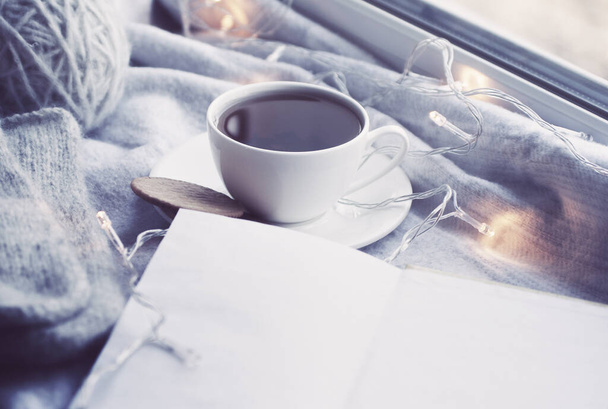 Cozy winter still life: mug of hot tea and book with warm plaid on windowsill against snow landscape from outside. - Fotoğraf, Görsel