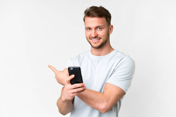 Young handsome caucasian man over isolated white background using mobile phone and pointing back - Fotografie, Obrázek