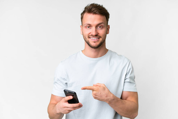 Young handsome caucasian man over isolated white background using mobile phone and pointing it - Foto, immagini