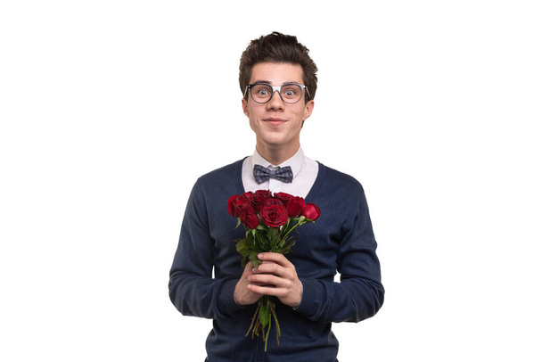 Glad young male in smart casual clothes and glasses with bunch of red roses smiling and looking at camera during date against white background - Photo, image