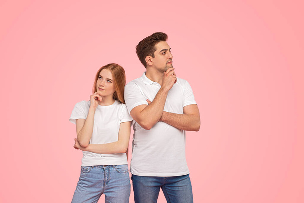 Pensive young man and woman in similar clothes touching chins and thinking, while standing near each other and looking in different directions against pink background - Photo, image