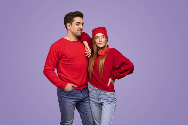 Young woman in stylish outfit with hat and headphones leaning on shoulder of boyfriend against violet background - Foto, imagen