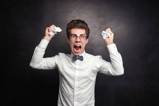 Aggressive young man in smart casual clothes and glasses tearing and clumping paper while screaming in anger against black background - Foto, immagini