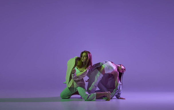 Studio footage of dancing young girls. Stylish performance. Two young women in expressive contemporary dance isolated over purple background. New dance style, youth, music and fashion, ad, trends. - Fotografie, Obrázek