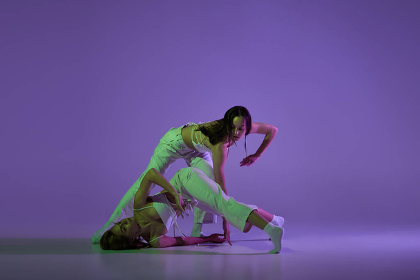 Experimental dance. Two young girl in motion, action isolated over purple background. Concept of new dance style, youth culture, music and fashion, ad, trends. Flexible and graceful dancers - Photo, image