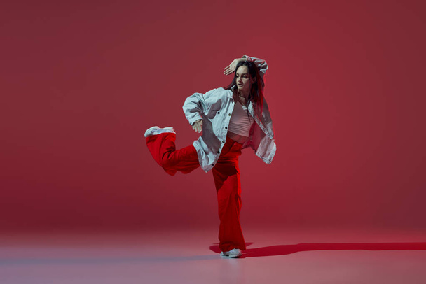Retrato dinámico de una joven bailando hip-hop o danza contemporánea aislada sobre fondo rojo oscuro. Danza experimental. Música, danza, estilo de vida activo, moda, estilo. Energía y flexibilidad - Foto, imagen
