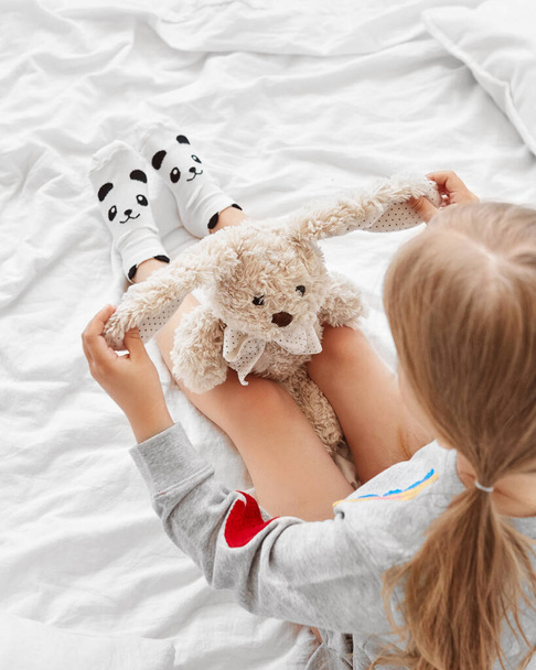 Vue du dessus de la fille tenant peluche lapin jouet assis sur le lit portant des chaussettes panda mignon - Photo, image