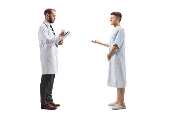 Full length profile shot of a male doctor and a young male patient in a hospital gown standing and talking isolated on white background - Fotó, kép