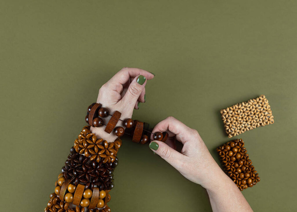 Kobieta zakłada na ręce bransoletki wykonane z ręcznie robionych drewnianych koralików brązowo-beżowych na zielonym tle. Styl boho, retro hipisowska dekoracja. Widok z góry - Zdjęcie, obraz