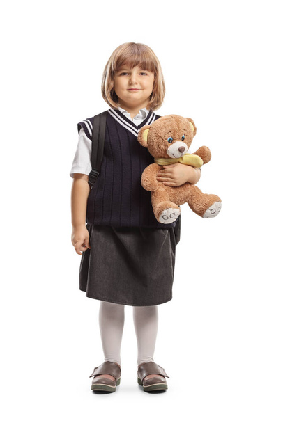 Full length profile shot of a little schoolgirl in a uniform holding a teddy bear isolated on white background - Foto, Imagem