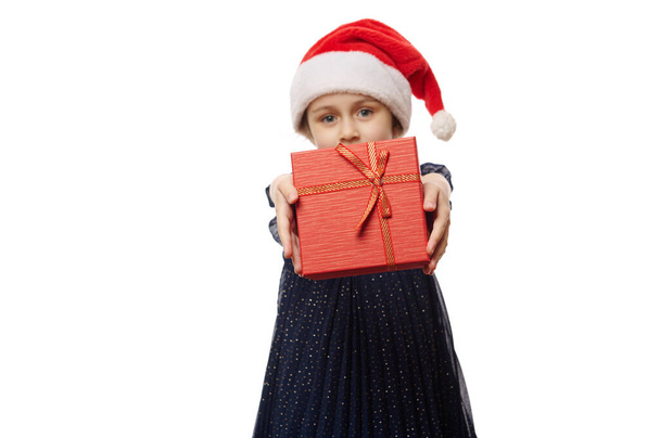 Szelektív összpontosítani egy elegáns piros ajándék doboz kezében egy aranyos kislány, tartja a karácsonyi ajándékot a kamera, elszigetelt fehér háttérrel. Boksz nap. Karácsonyi és újévi ünnepség koncepció - Fotó, kép