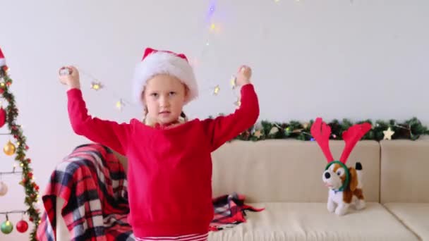 Smiling child girl in Santa Claus hat decorates herself with Christmas lights for CELEBRATED Christmas . Portrait of Christmas girl. - Footage, Video