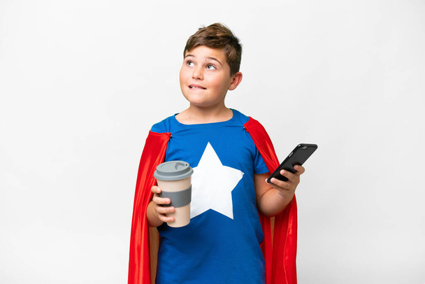Хлопчик-супергерой над ізольованим білим фоном, тримаючи каву, щоб забрати її і мобільний, думаючи щось - Фото, зображення