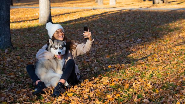 Nuori nainen halaa huskyaan ja ottaa selfien älypuhelimella. Nykyaikaiset trendit sosiaalisissa verkostoissa ja mahtaileva rakkaus eläimiin. - Valokuva, kuva