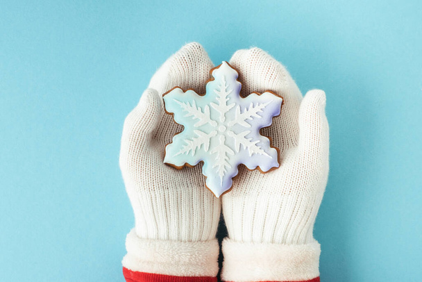 Woman hands wearing white mittens and holding snowflake shaped gingerbread cookie with festive icing on the blue pastel background. Christmas background - Фото, изображение