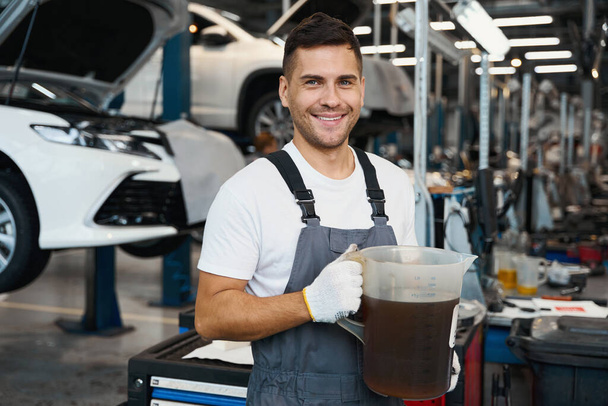 Portrét mechanika ve speciálním obleku stojící v garáži, držící džbán se strojovým olejem - Fotografie, Obrázek
