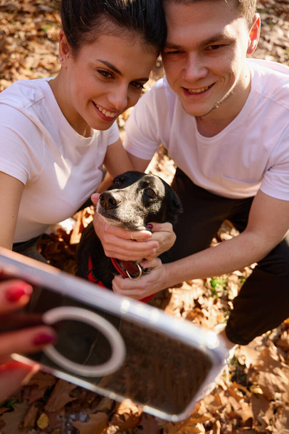 Felice coppia di studenti prendere selfie con il loro cane sullo sfondo della natura autunno, sorridono alla fotocamera del telefono - Foto, immagini
