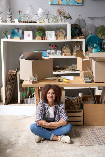 Full length portrait of smiling young curly female sitting on floor near cardgoard boxes and gifts for home - Photo, Image
