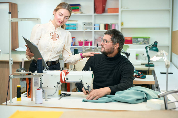 Mooie vrouw staat aan tafel met een naaimachine, een man zit op het werk - Foto, afbeelding