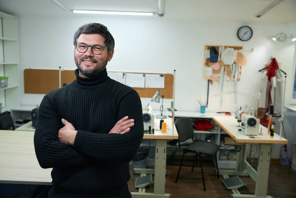Un tailleur souriant se tient dans un atelier de couture au milieu de la pièce, un maître en lunettes et pull tricoté - Photo, image