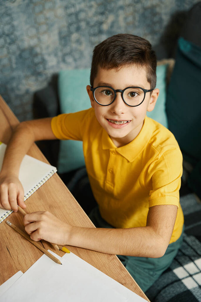 Portrét dítěte, jak sedí u stolu, drží tužku a doma dělá domácí úkoly, dívá se do kamery - Fotografie, Obrázek