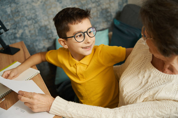 Ребенок сидит за столом, смотрит на бабушку, держит бумагу, делает домашнее задание - Фото, изображение