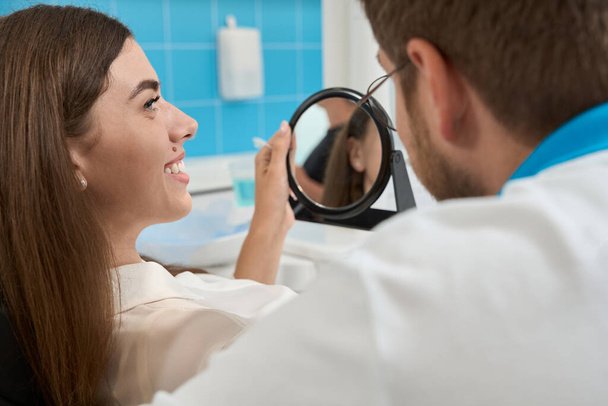 Pleased female patient seated in chair holding hand mirror and looking at her dentist - Foto, immagini