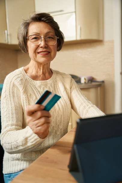 Alte Frau mit Brille sitzt am Tisch mit Tablet, hält Kreditkarte in der Hand und schaut auf Papier - Foto, Bild