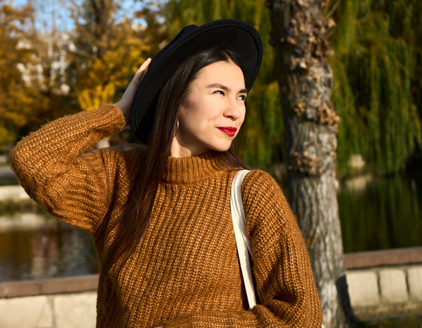 Selective focus of smiling fashionable young tanned woman in hat among autumn foliage outdoors in city park autumn. Positive emotion concept. High quality photo - Foto, Imagem