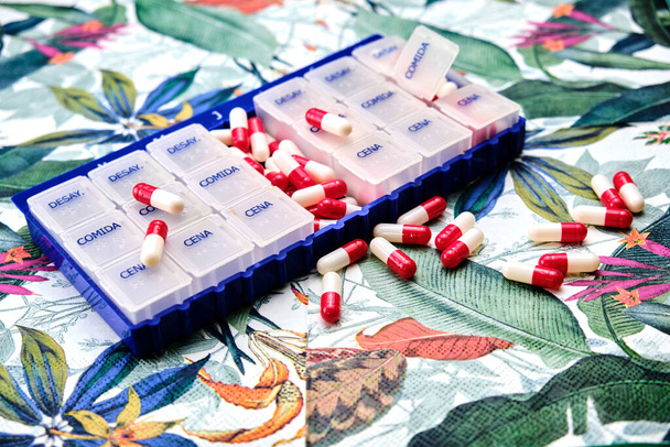 A hét napjai pillérdoboz tele piros-fehér tabletták tetején egy asztal egy trópusi minta. - Fotó, kép