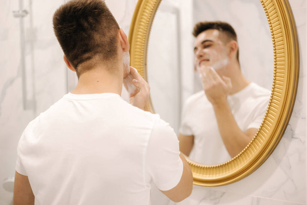 Handsome man washing face with facial cleanser. Face wash soap in bathroom sink at home. Male in white t-shirt. - Foto, afbeelding