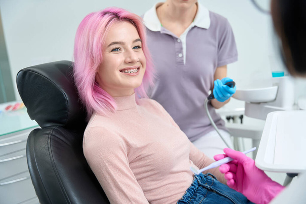 Розововолосая улыбающаяся женщина с брекетами на приеме у стоматолога, врач и ассистент используют специальные инструменты - Фото, изображение