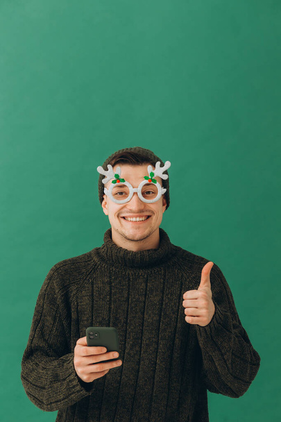 A young man in a warm sweater, carnival glasses and a phone in his hands, isolated on a green background. - Photo, Image