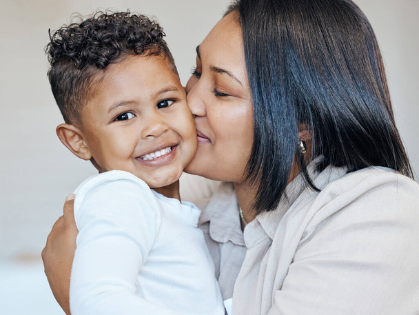 Mujer de raza mixta besando a su adorable hijito en la mejilla mientras se unen en casa. Pequeño niño hispano sonriendo y mirando feliz de estar recibiendo amor y afecto de su madre. - Foto, Imagen
