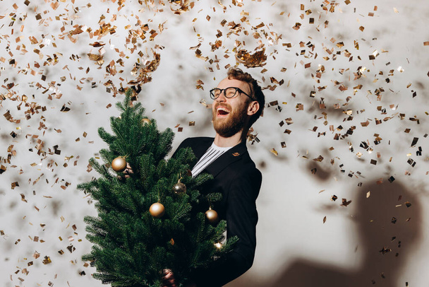 Roodharige knappe man in een zwart jasje en een bril danst met een kerstboom in zijn handen. Nieuwjaarsfeestconcept. - Foto, afbeelding