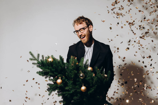 Červenovlasý pohledný muž v černém saku a brýlích tančí s vánočním stromečkem v ruce. Novoroční párty koncept. - Fotografie, Obrázek
