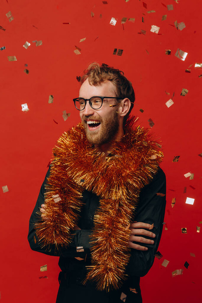 Homme joyeux aux cheveux roux dans des verres et un boa doré à la fête du Nouvel An avec des confettis. Studio photo. - Photo, image